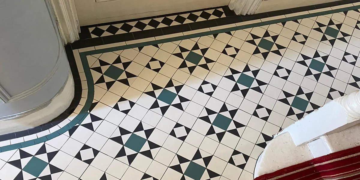 Looking down at a Georgian-style mosaic tiled hallway floor as the tiles are cut to curve around the wall corner. The floot of the white painted staircase is to the right, with a deep red striped stair-runner.