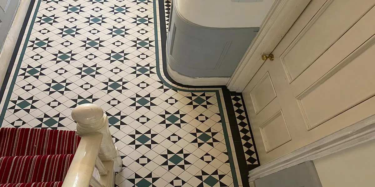 A view from the top of the staircase, looking down on a hall floor with a black, white and green mosaic tile pattern. The door thresholds have a row of chequered tiling. The gloss-white staircase has a burgundy stair-runner.