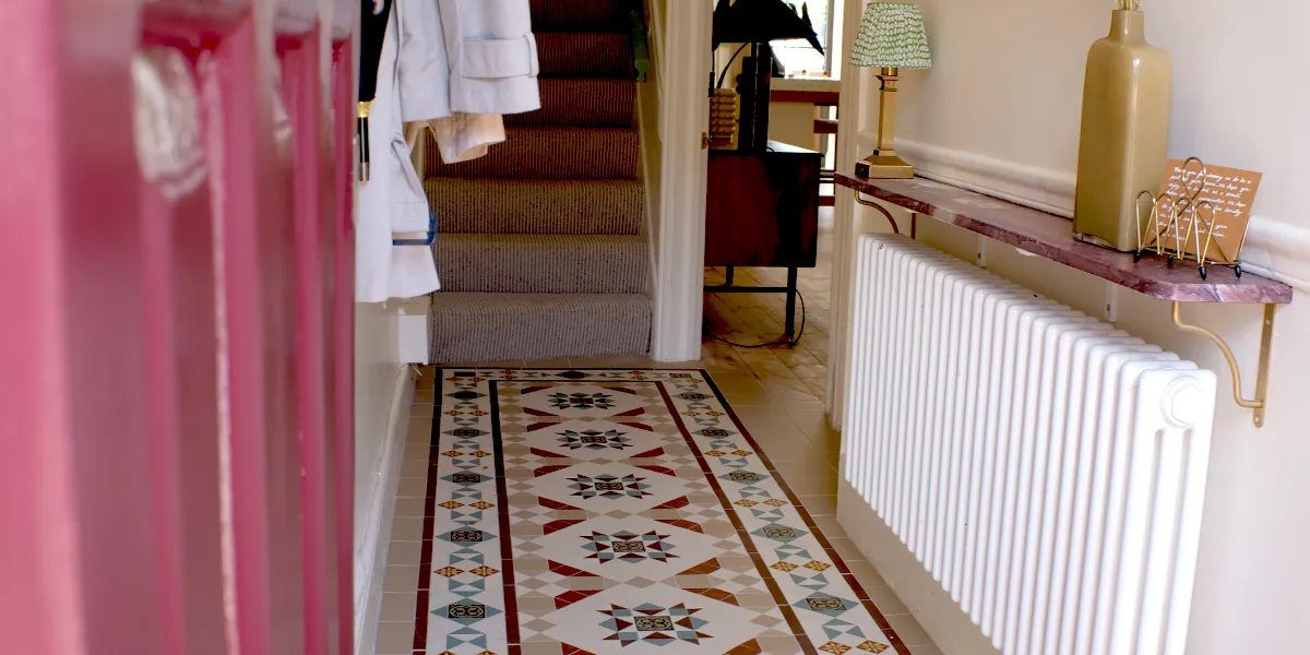
	A highly decorative mosaic in a Victorian rug style leads through a narrow hallway to a staircase with grey carpet. To the left a pink-painted front door is open. To the right a marble shelf sits above a white-painted radiator.