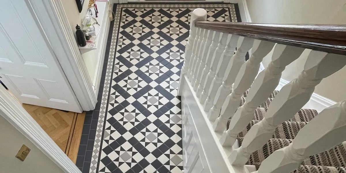 View looking down at a high-contrast black, white and grey Victorian style tiled floor from halfway up a staircase. The mosaic pattern combines star and box motifs, neatly framed by a grey and white diamond border. The doors, skirting, staircase and its spindles are painted gloss-white. The bannister is dark wood, and the staircase has a patterned stair-runner. To the left, an adjoining room has parquet floor.