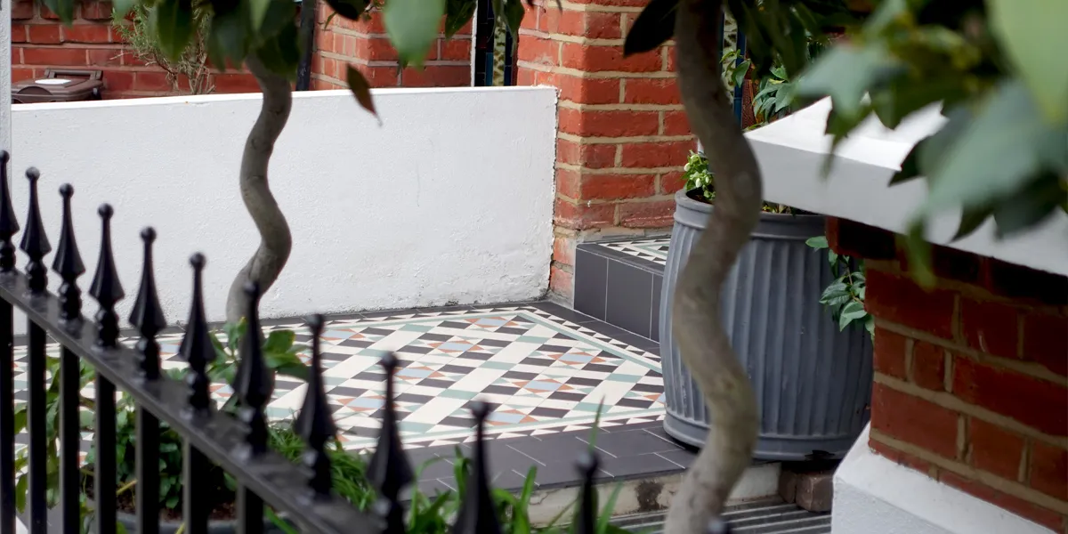 A view through an out-of-focus iron-railing fence and twisting tree trunks towards a Victorian tiled path. The colourful pattern uses black, white, red, blue and green colours. The house is red brick, with white rendered base, walls, and window sills.