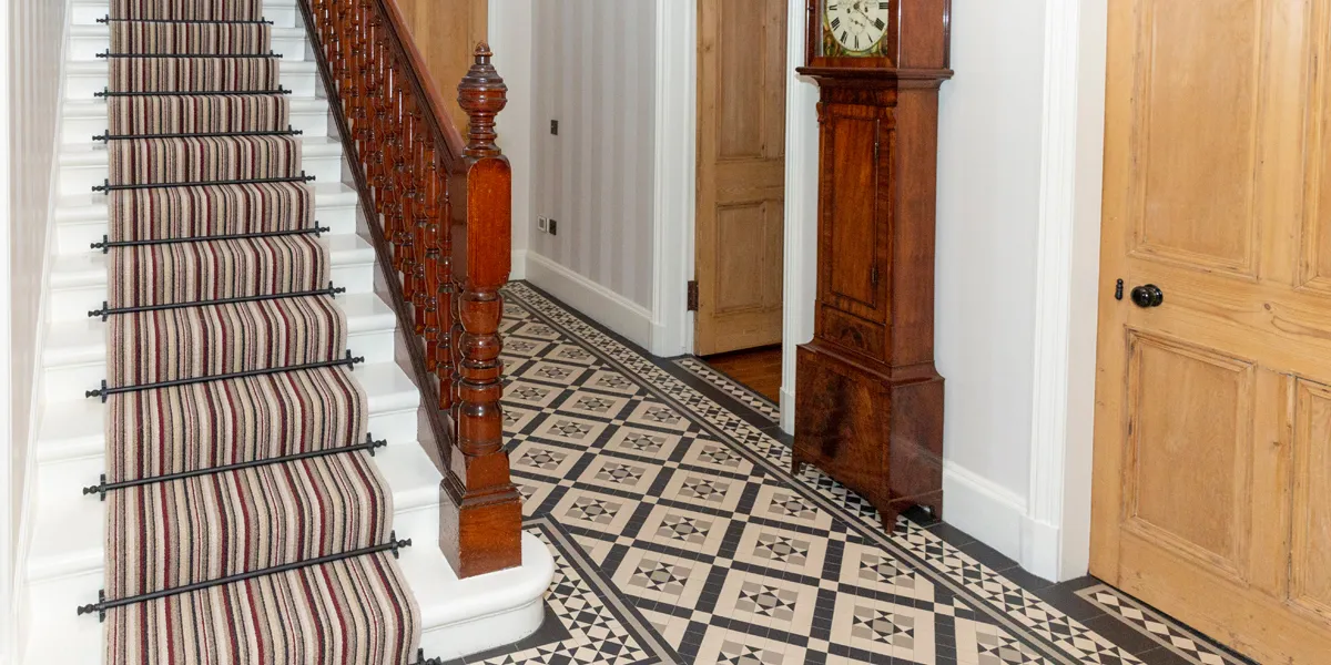 A decorative geometric tiled floor in a hallway. The staircase has a multi-coloured carpet runner and ornate wooden banister and spindles. To the right, an antique grandfather clock sits between two wooden doors leading to other rooms in the house.