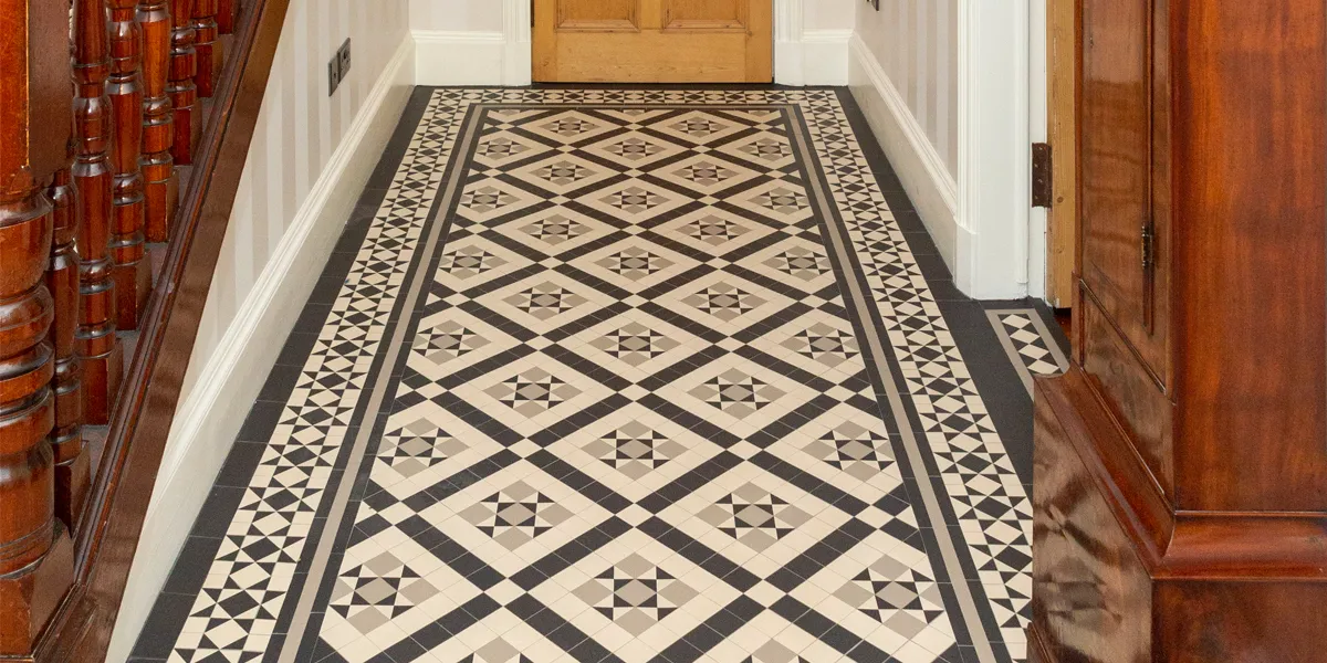 Black, white and pale grey mosaic hall floor tiles lead towards a wooden door along the side of a staircase with ornate woodwork. To the right, there is the base of an antique grandfather clock. There is subtle striped wall paper, and gloss white skirting and architraves.