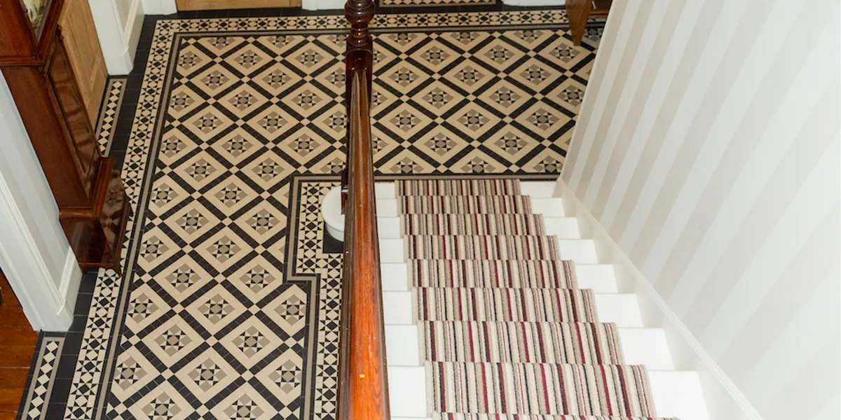 View from the top of a staircase looking down towards a decorative geometric tiled floor in the Victorian-style. A wide border motif follows the perimeter of the space. The staircase treads are painted gloss white, with a colourful striped stair-runner. To the left, a grandfather clock between two door thresholds.