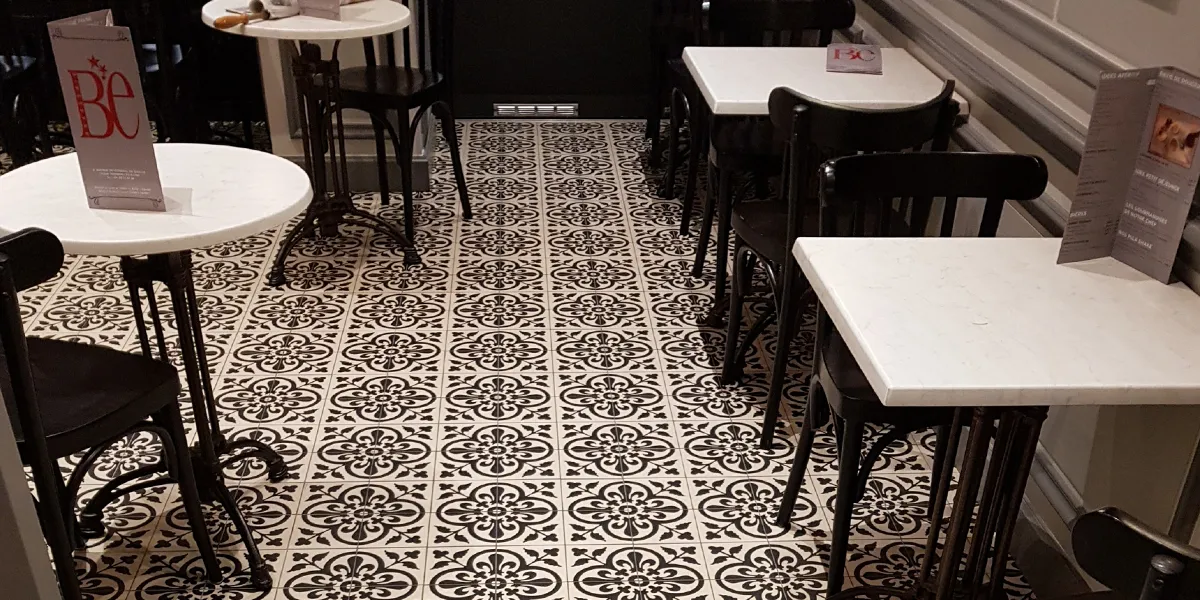 Art-nouveau inspired European style encaustic tiles on cafe floor. White marble table-tops with black metal legs, and dark-wood chairs. The cafe menu sits on top of the tables.