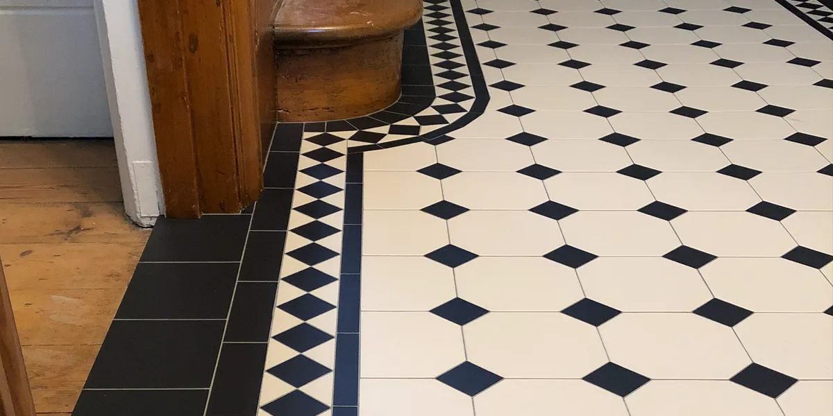 Close view of Victorian octagon and dot tiles in a hallway. The border pattern curves around the foor of the wooden staircase. A open doorway to the left reveals wooden floorboards.