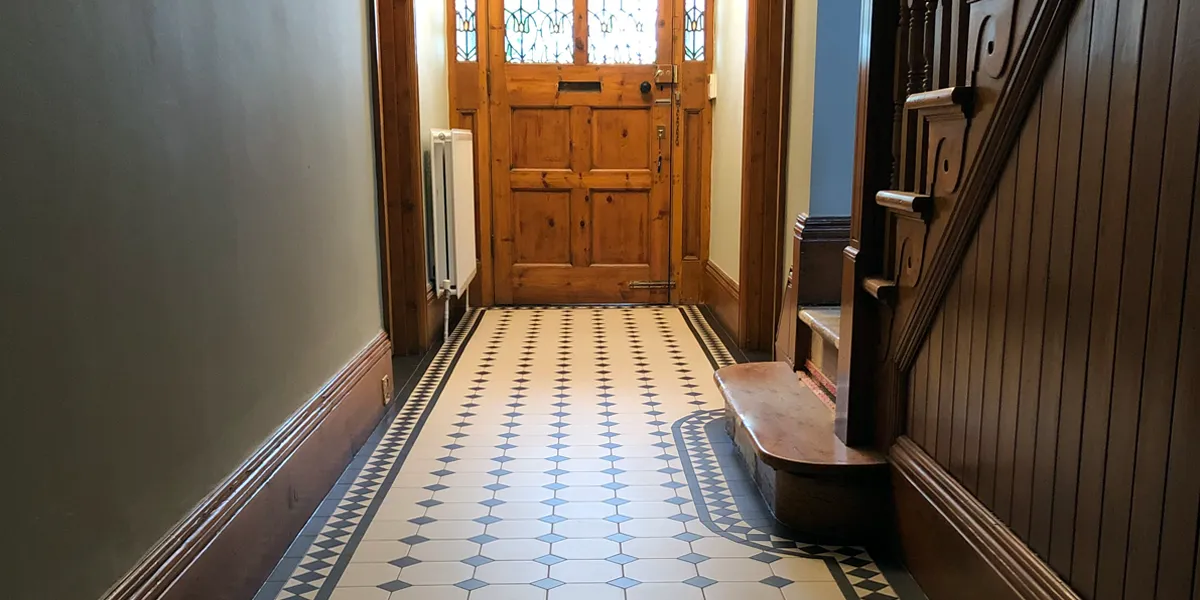 View from the back of a hallway towards a wooden front door with stained glass, which allows light to illuminate the middle of the floor. The floor is comprised of Victorian octagon tiles. The staircase, rises to the right and has simple panelling to the side. All the woodwork is natural and varnished with a dark oak stain.
