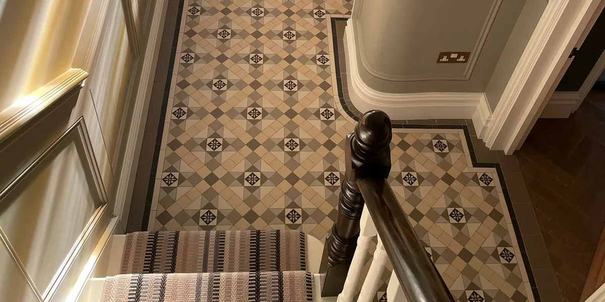 Looking down from a hallway staircase at a detailed Victorian-style mosaic floor in grey and beige tones. The mosaic design includes a decorative encaustic tile in the centre of the motifs, and is framed with a simple three line border. The staircase has a striped stair-runner. The skirting and architraves are painted white, while the walls have wooden panelling painted a soft grey. A diffuse warm light is seen on the wall to the left, and the staircase newl post and banister are dark stained wood.