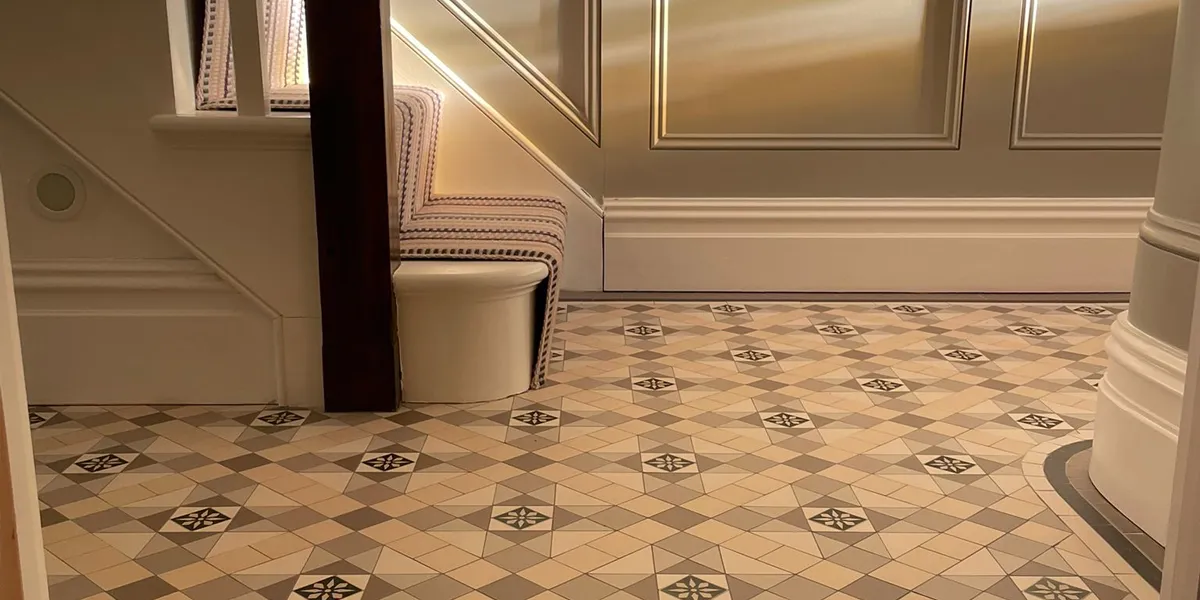 Side-view of a mosaic tiled hallway floor and base of staircase. The staircase and skirting are painted white, and the wooden panelling a light-grey colour. Light is seen falling on the rear wall, also lightening the striped stair-runner.