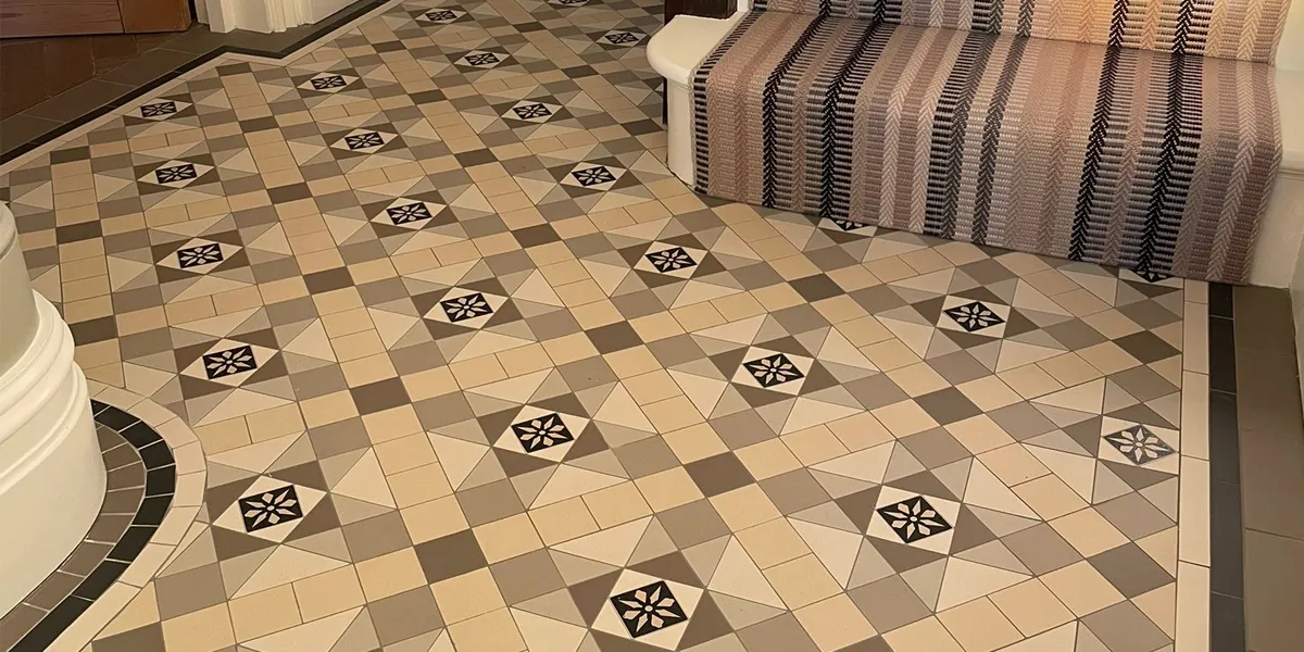 Close view of Victorian mosaic tile design at the foot of a staircase in a hallway. The mosaic tiles are warm greys and beige tones. The staircase has a striped stair runner which complements the colour of the mosaic tiles.