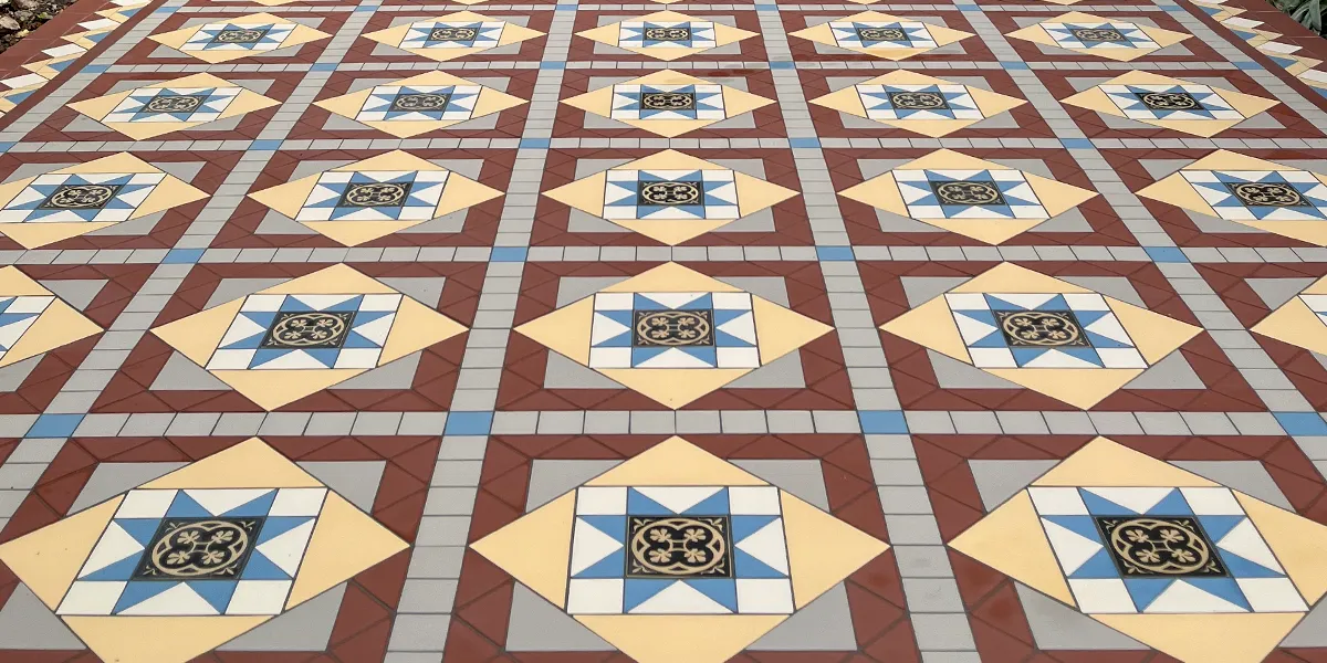 Close-up view of a colourful mosaic tile pattern. Mainly composed of a grid of small grey squares. Red, yellow and blue triangles surround an encaustic tile at the centre of each motif.