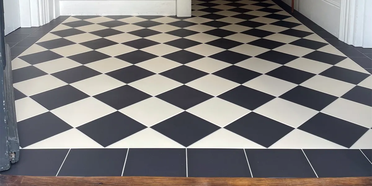 Black and white hall tiles viewed from floor level at the front door. The foot of the staircase and skirting-boards can be seen to the top and right.
