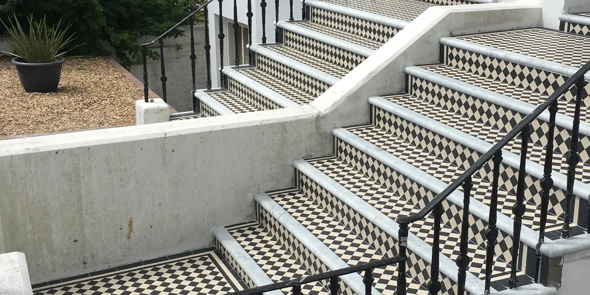 View across two sets of steps, which are finished with Victorian style small black and white chequered tiling. The front edge of the steps has a marble tread, and the steps are lined with black metal railings. A rendered wall with chamfered edge seperates the two sets of steps. To the rear we see a front garden area covered in gravel, with a black pot containing a spikey green plant.