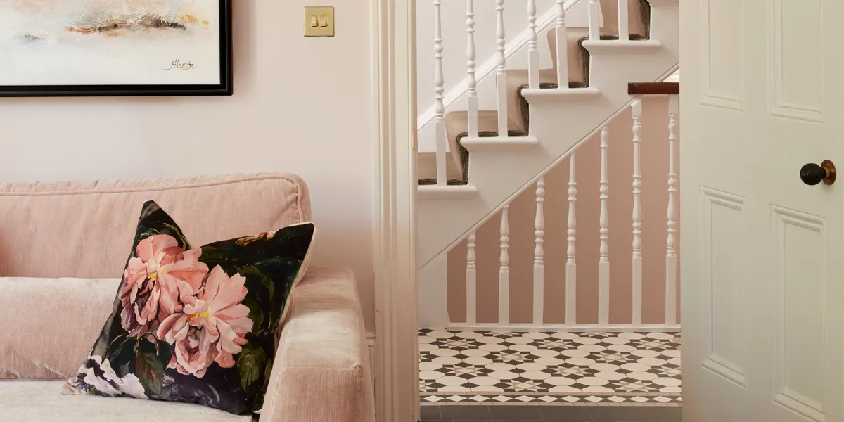 View through a living-room door towards the hallway and staircase rising. In the hallway floor a Victorian mosaic design. The livingroom has a pale-peach sofa, with a cushion printed with large pink flowers.