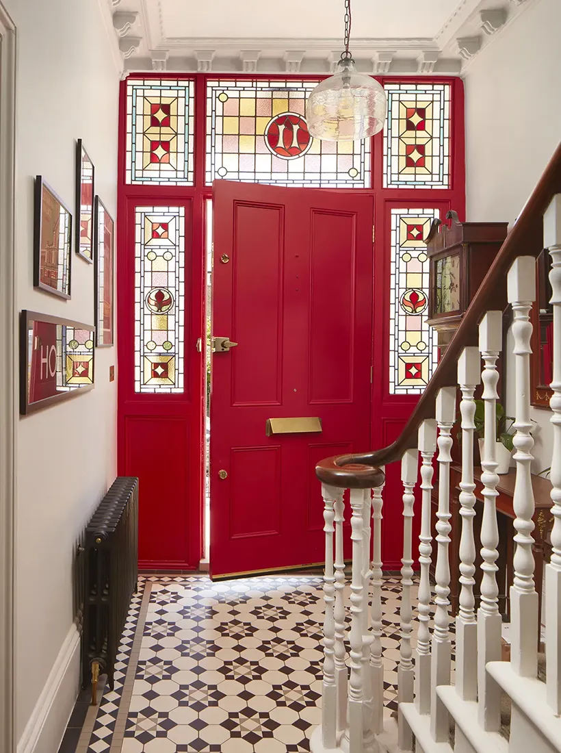 A traditional black and white Victorian mosaic hall floor design using octagons contrasted with a star motif. A large red front door is ajar and natural light reflects off the tiles beneath. To the right the spindles and curved banister rail of a staircase rise. On the left, a black antique-style radiator.