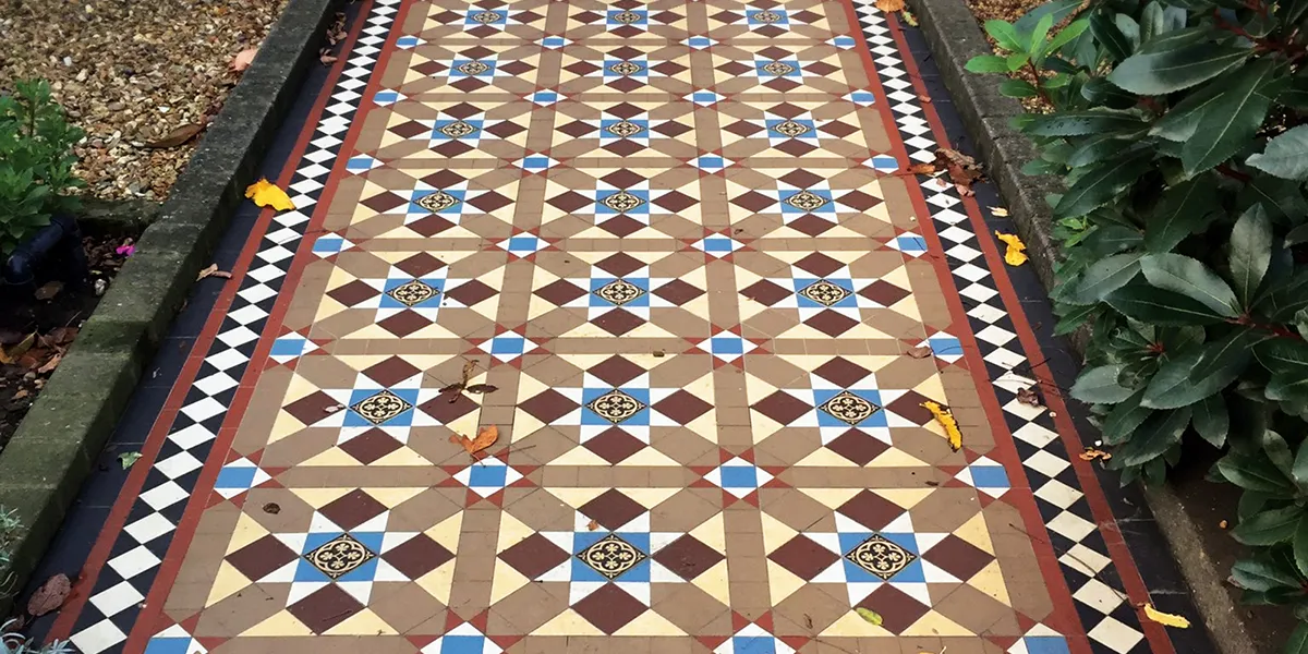 A multi-colour Victorian encaustic tile path with some autumn leaves fallen on it. The sides of the path are old stone setts. To the left an area of gravel, to the right a deep green bush.