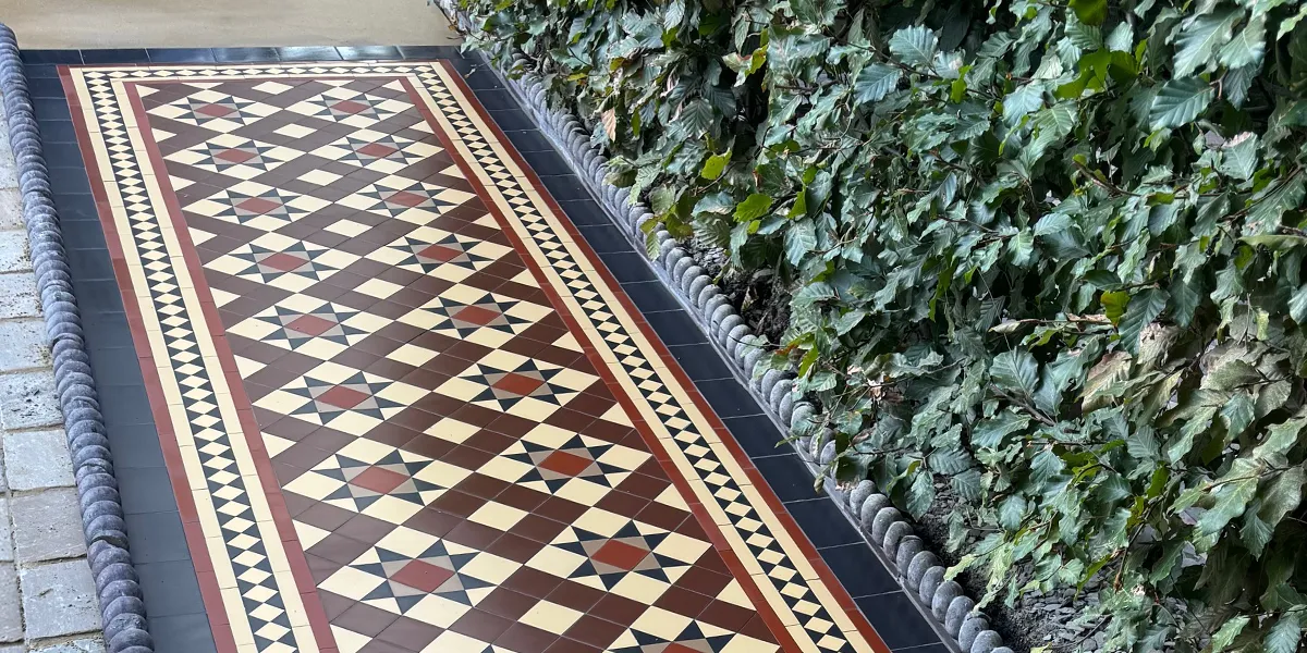 An Edwardian-style front garden path in an earthy colour palette. To the right a beech-tree hedge, and the rest of the garden has stone setts. At the top of the path, a stone threshold is fitted flush with the tiling.