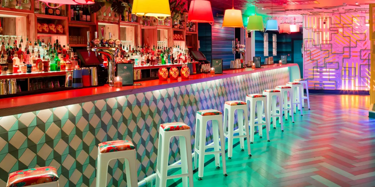 A modern bar scene with multi-colour lighting. The front of the bar has a geometric tile pattern in pink, blue and grey colours, forming a 3D cube motif. In front of the bar there is a row of white plastic stools. Behind the bar there hundreds of bottles of spirits and other types of drink. The floor of the bar is a herringbone tile pattern.