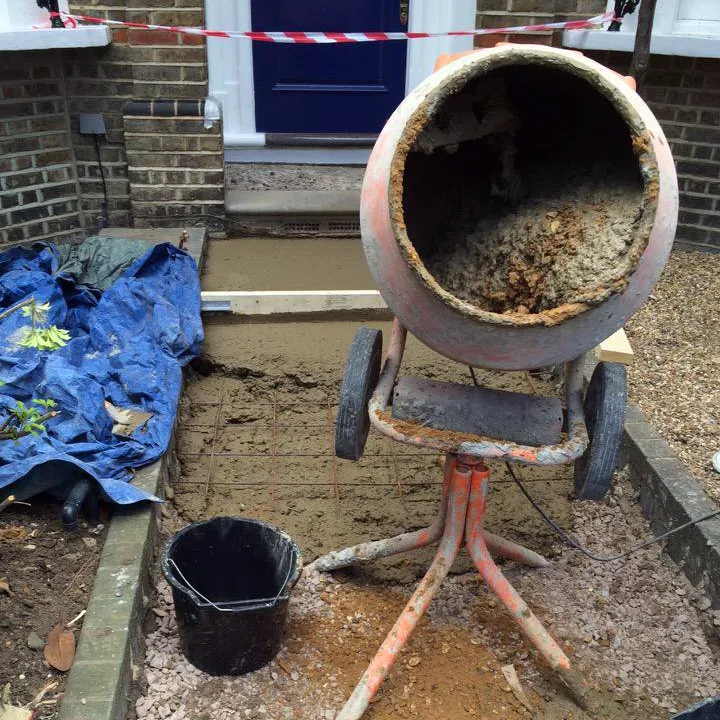 A sharp-sand and cement pathway base is being prepared. Steel mesh is seen within the cement. To the right there are old blue-coloured plastic sheets, and to the front a dirty and well-used cement mixer and bucket.