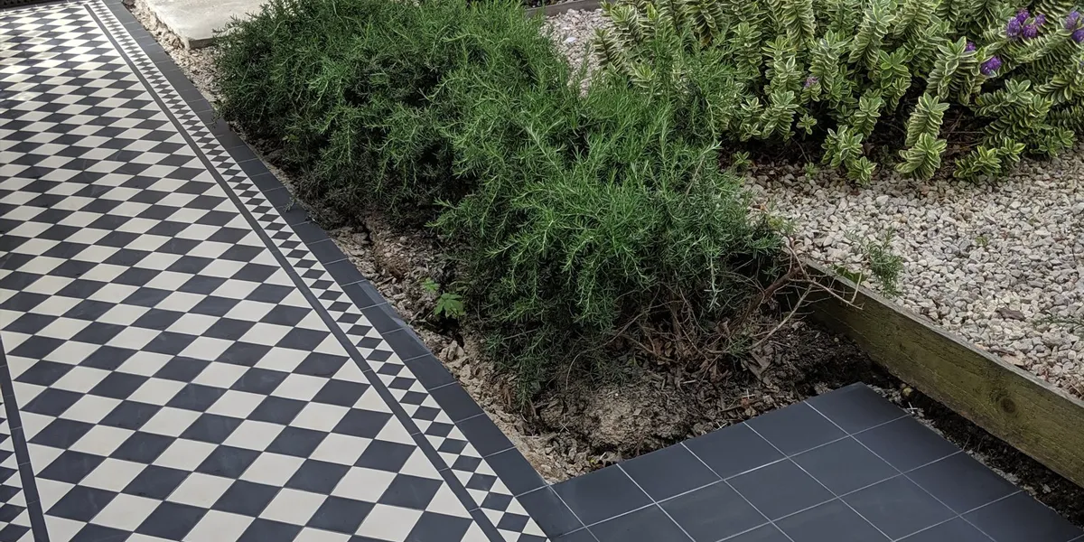 Black and white chequerboard path tiles to the left, with a garden to the left with lots of wild shrubs between pebbles.