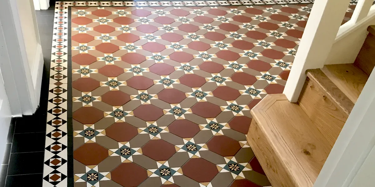 A detailed, mainly red toned, mosaic tile hallway floor featuring black and white encaustics at the intersection of the pattern. The border mosaic contrasts in white, black and red tones. To the right, a wooden staircase rises, with gloss white newel post and sides.