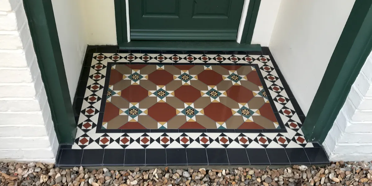 A porch with an Edwardian-style tiled floor. The detailed pattern features red octagons, and an encaustic at the centre of each motif. The border mosaic uses a contrasting black, white and red diamond design. The porch is framed by two deep-green painted columns, matching the doorway. The remainder of the walls are painted white. The porch steps down to a gravel path.