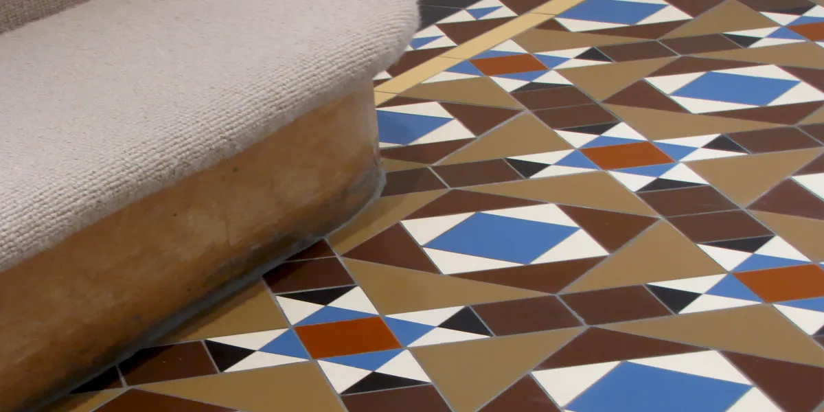 Close up view of Victorian style hallway floor tiles around the base of a staircase. The mosaic pattern is composed of different sized and coloured, squares and triangles. The staircase tread has a beige carpet fitted.