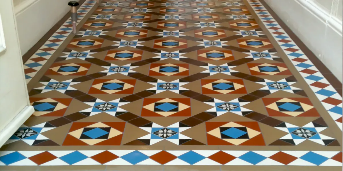 Traditional multicolour Victorian hallway tiles featuring a black and white floral design encaustic. Red, blue and brown tones dominate this Victorian floor tile design. The hallway has simple white skirting boards, and to the left we see the edge of the open front door, also painted white.