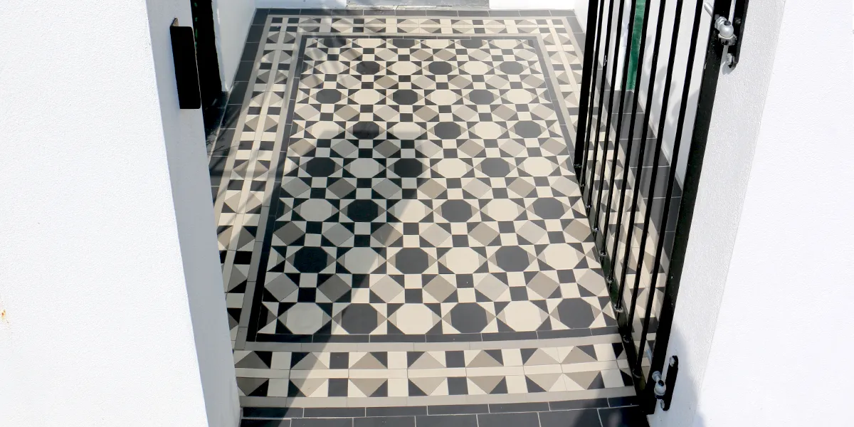 View between two white rendered gate posts of a modern mosaic path design. The tile pattern has a 1960s style, made of octagons, squares and triangles, in black, white and grey tones. To the right, a simple black metal gate hangs open.
