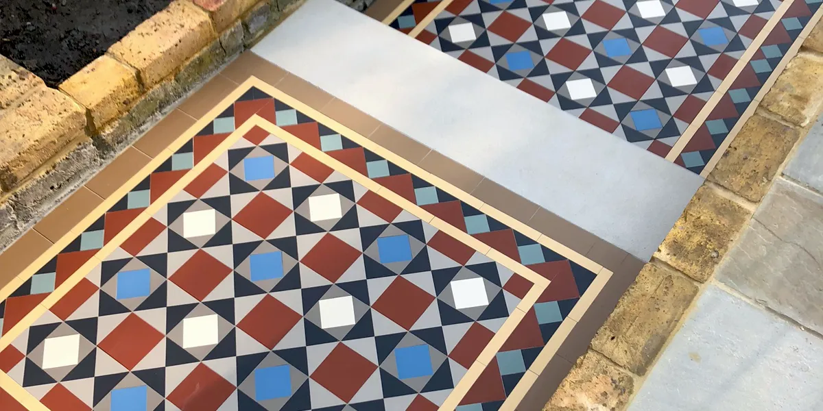 Looking down at a Victorian mosaic pathway, with a stone step tread in the middle. The mosaic is multi-coloured, but predominantly uses red tiles. To the edges of the path, a yellow brick wall retains the garden. Soil for planting on the left, and stone slabs to the right.