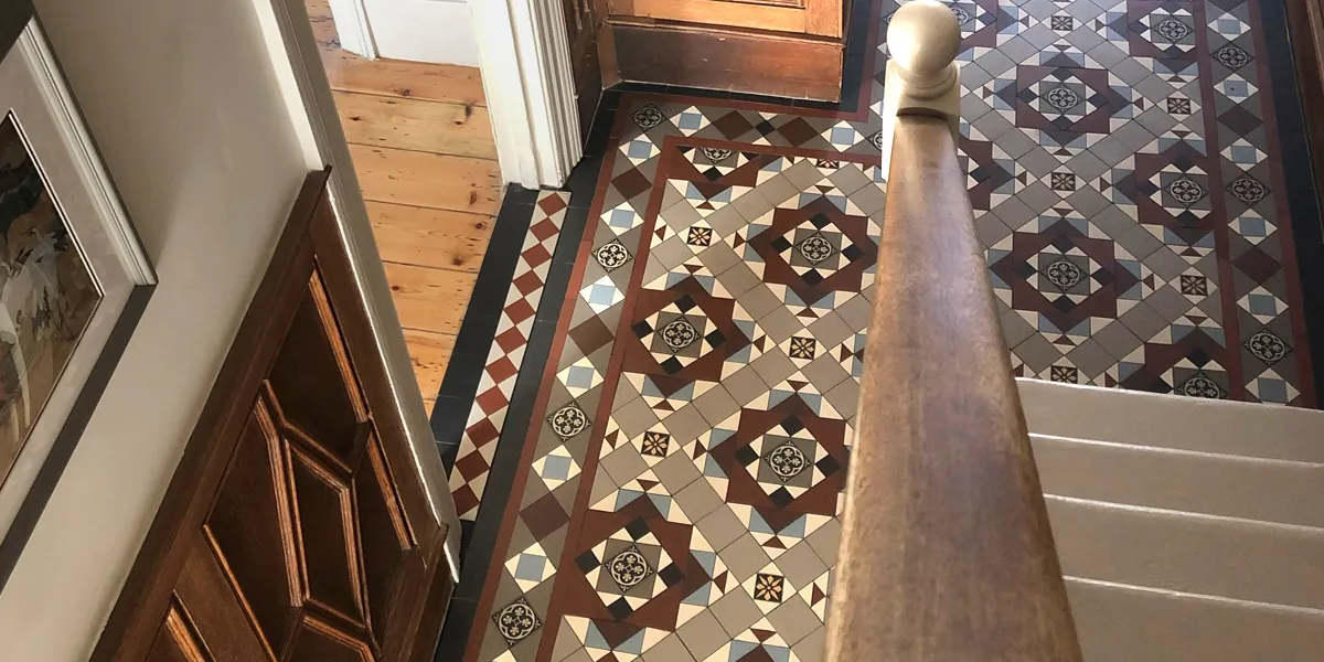 Looking down from a hallway staircase at a Victorian encaustic floor tile mosaic below. The staircase treads are painted gloss white, and the handrail is wood. To the left we see a threshold through to another room, where a red diamond motif border mosaic is fitted between the door architraves. The adjoining room has exposed wooden floorboards. The wall to the left has wooden panelling, and above this a picture hangs.