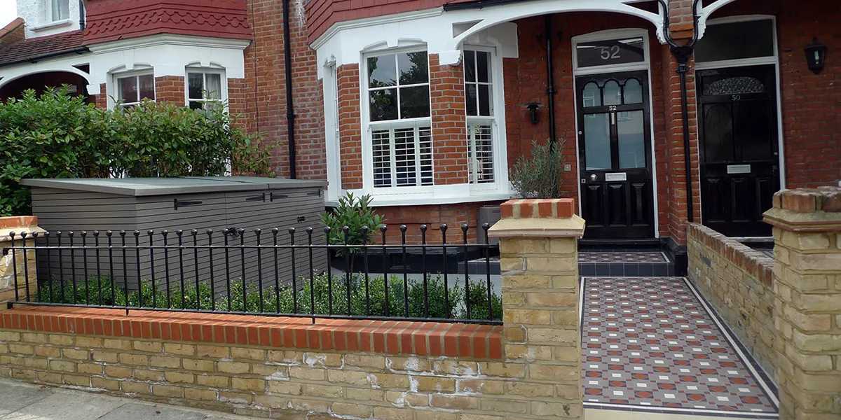View across the front garden of a property. The front garden walls are made of London-yellow brick, topped with contrasting red bricks, and iron railings. An Edwardian style mosaic path leads fron the pavement, to a raised porch and black gloss painted front door. Behind the low garden wall, there are green shrubs, and to the left a wooden bicycle store in front of the boundry hedge. The house is red brick with a bay window, which has white painted stonework