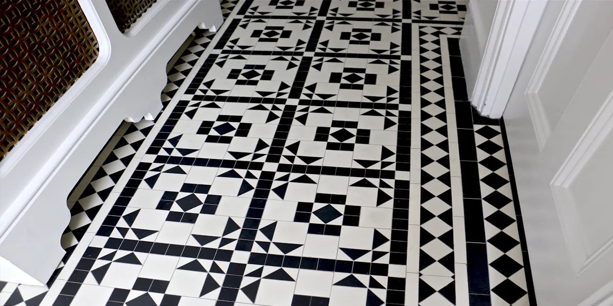 Close up view of a highly detailed black and white mosaic floor tile design in an entrance hall. To the left we see the base of a traditional radiator cover. To the right a gloss-white painted door.