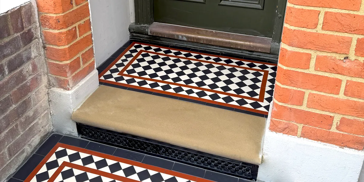 View of a porch step with a York stone tread leading to an area of Victorian style black and white tiles. The design uses red lines in the border. Beneath the stone tread, a daisy grate ventilation grill is painted black. The house has red bricks, and white render at the base.