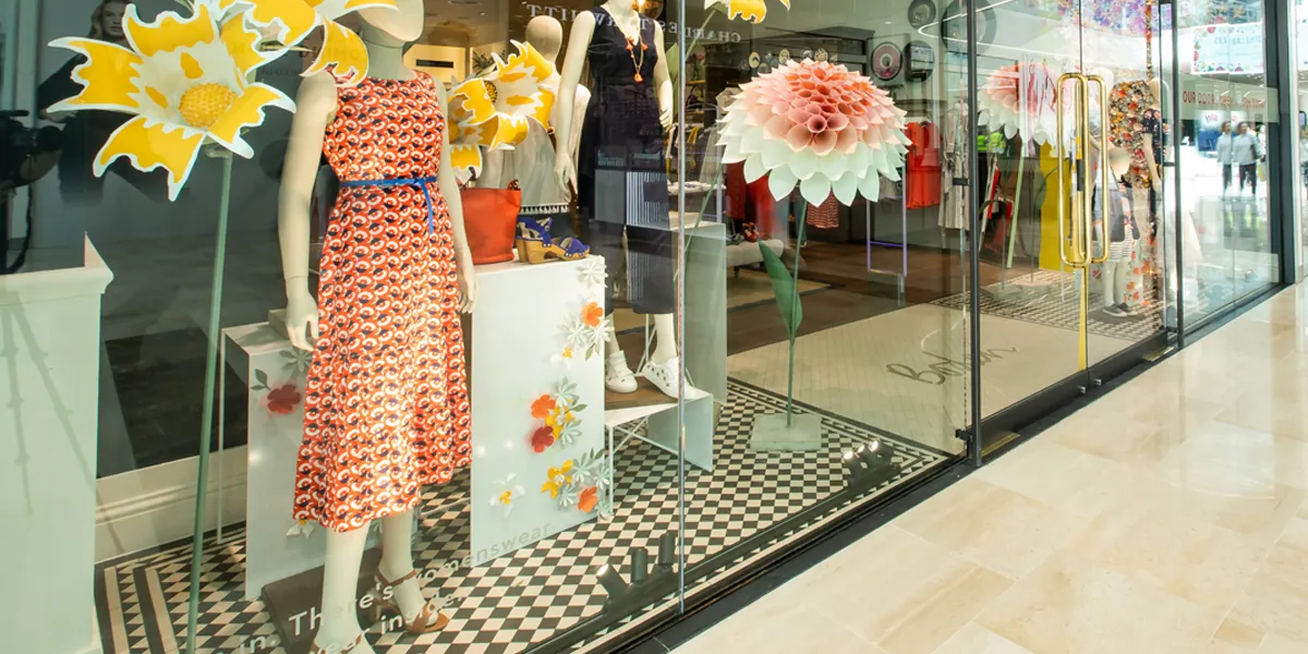 The front window display of a Boden shop in a shopping centre. Several dresses are on display, with large flower models either side. The floor of the window display is black and white chequerboard tiling.
