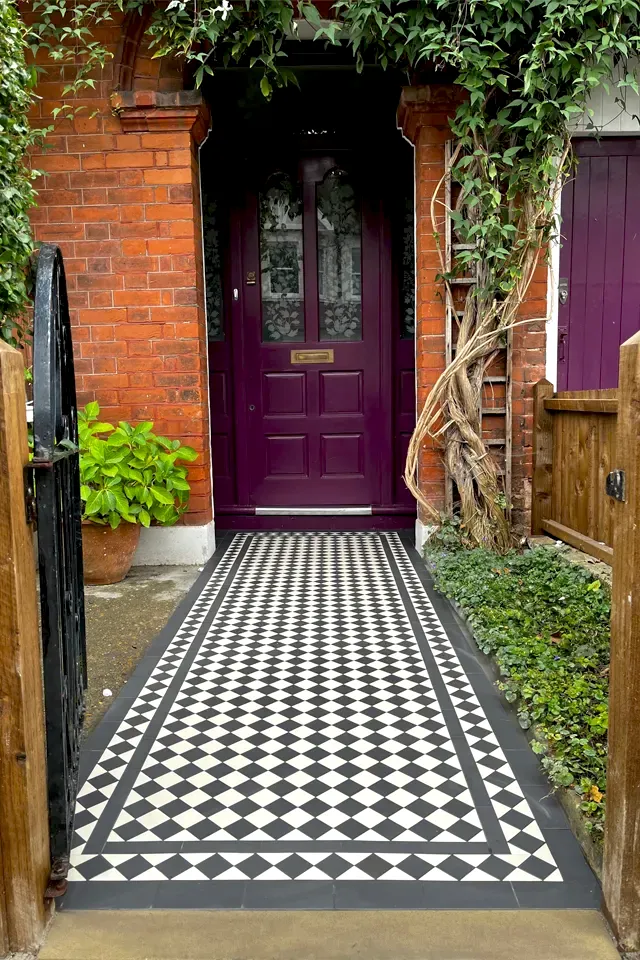 A traditional, Victorian style black and white chequered path leads to a purple front door, inside a small porch area. The facade of the house is weathered red bricks. To the left a potted plant with bright green leaves, and to the right, a twisting wisteria grows. On the right of the path, small shrubs and a wooden fence at the property boundry. At the start of the path, a York stone threshold is set between two wooden posts. To the left a black, wrought iron gate hangs open.