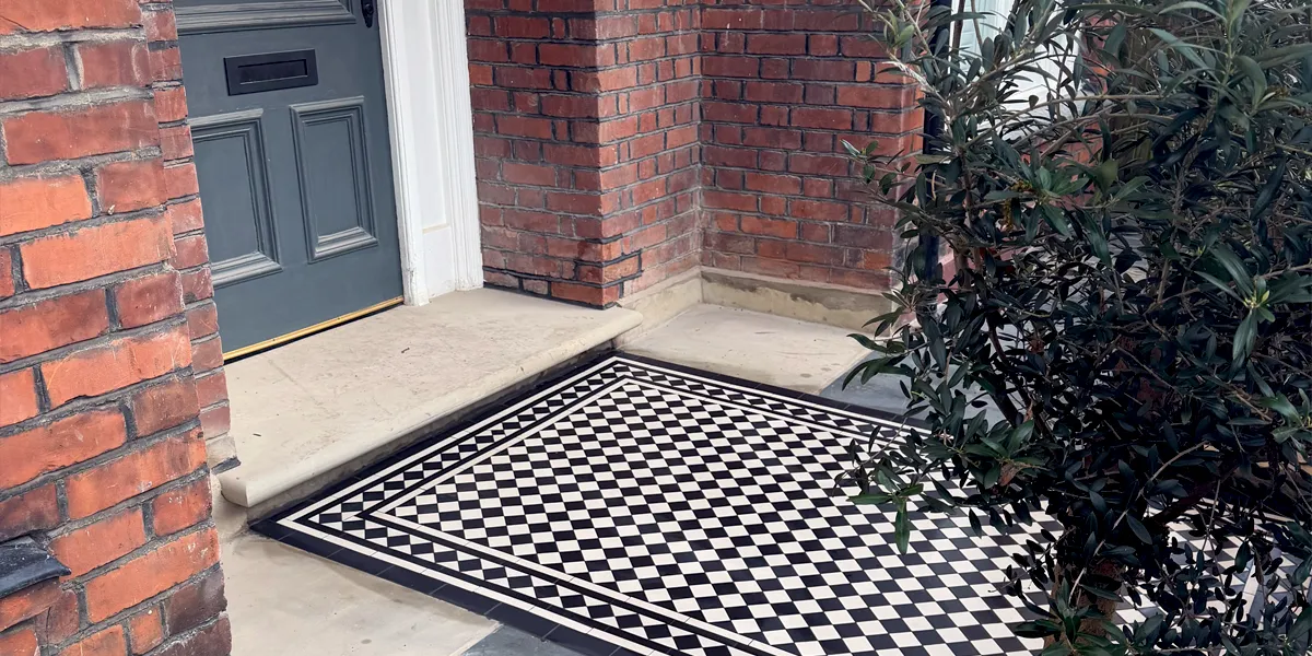 Side view of a Victorian style black and white chequerboard pathway leading to a large stone front door step. The property has a grey-blue front door, and old red bricks. To the right, a tall plant grows.