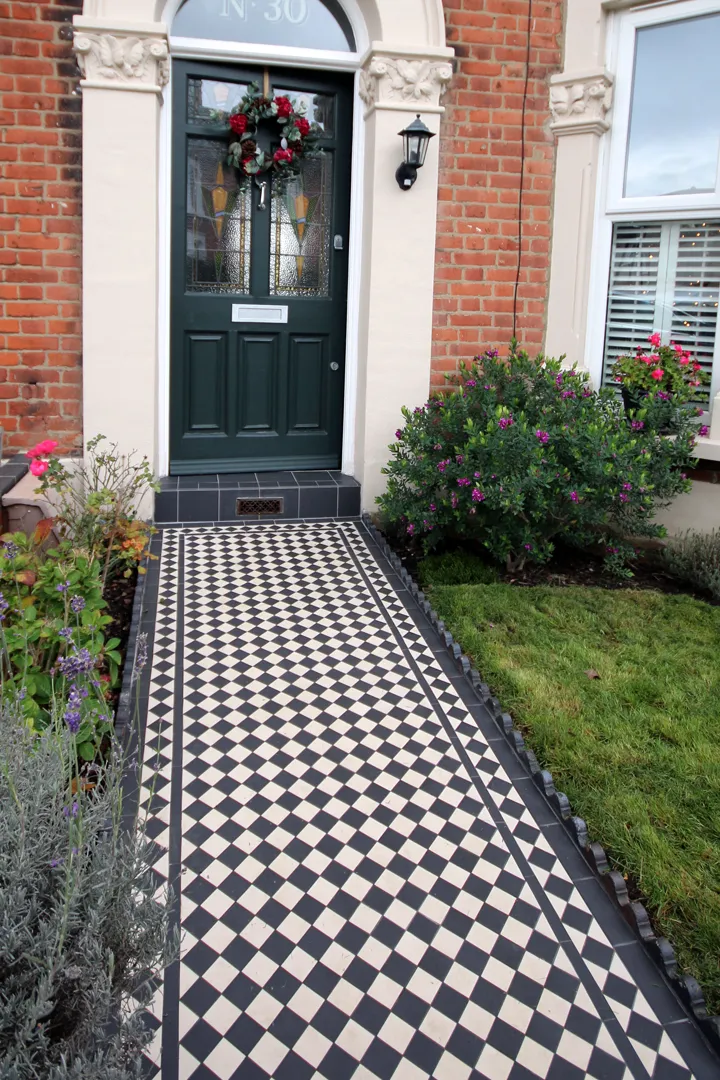 Traditional black and white garden pathway tiles, bordered with rope-top edging, leads to a large deep-green coloured front door with stained-glass inset. The red brick property has small shrubs, plants and flowers to the front and sides, and a small area of lawn.