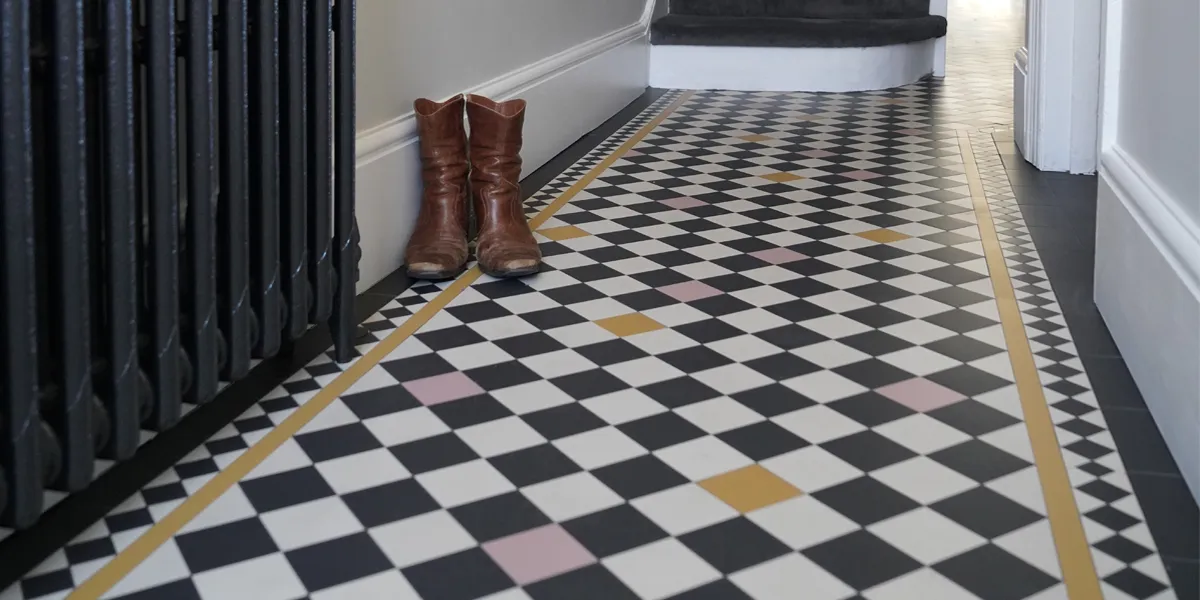 A traditional black and white mosaic hall floor with added splashes of pink and yellow tiles. To the left there is a black painted antique radiator and pair of brown leather boots.