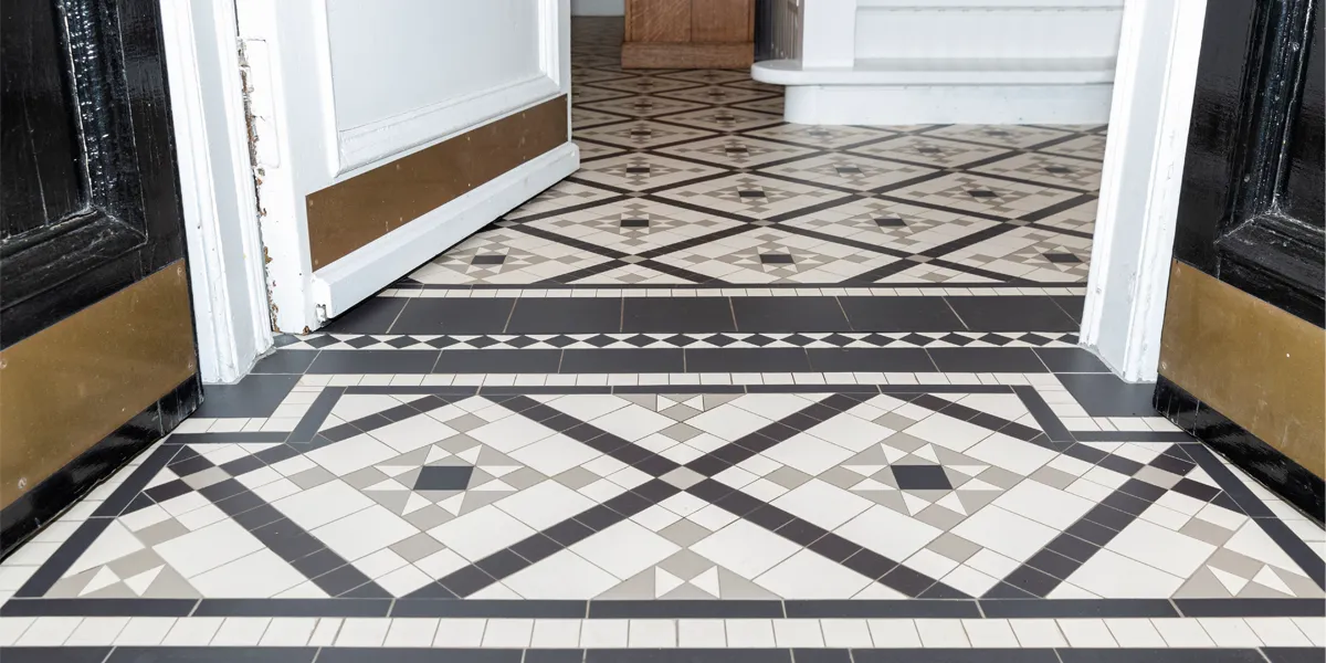 Low angled view through a vestibule towards a hallway. The floor is decorated with a detailed mosaic tile pattern in black, white and pale grey colours. The external vesibule doors are at the sides and painted black. THe internal door is painted white. All three doors have a brass plate at the base. In the hallway, the foot of a white painted staircase.