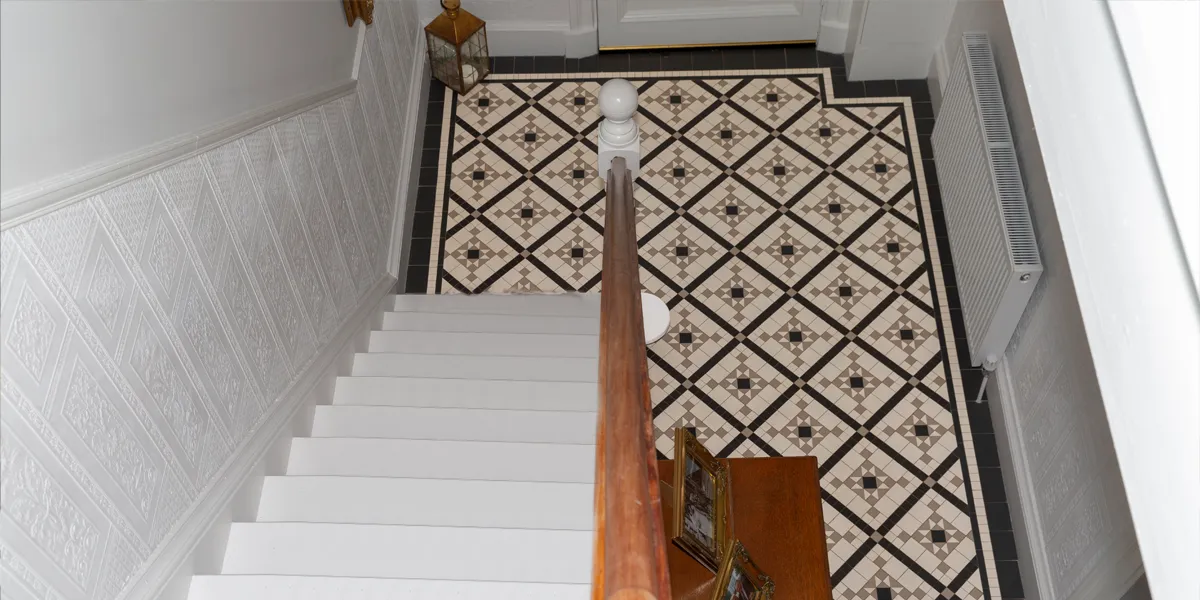 View looking from the top of a white painted staircase towards a Victorian-style of mosaic hallway floor. The tiles are black, white and grey, and the pattern has a trellis of black tiles with a grey square star motif between. The staircase has a wooden handrail, and the rest of the hallway is painted white.