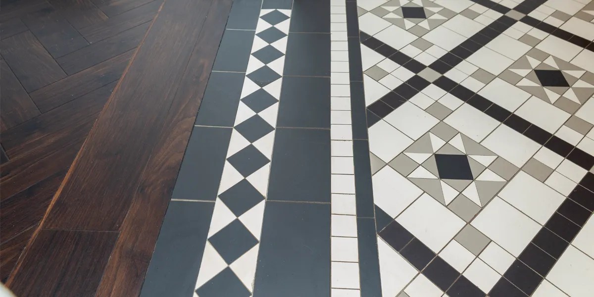 Close up view of some mosaic tiles in the space between a tiled hallway and adjoining room which has wooden flooring. The intricate and decorative mosaic pattern uses small square and triangles in black, white and grey colours.