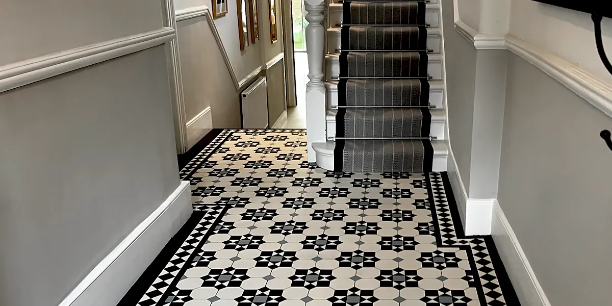 A hallway in neutral black, white and grey tones has a Victorian tiled floor leading to a white painted staircase with grey stripped stair runner. The hallway steps down to the rear, and the walls have white painted dado rails.
