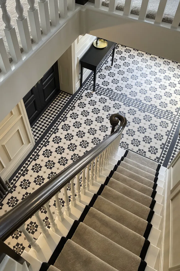 A view looking down a large staircase towards a grand entrance hall. The floor has decorative Victorian mosaic tiles, using a combination of octagons, squares and triangles in contrasting colours. Deep thresholds have a chequered pattern leading to large black doors.