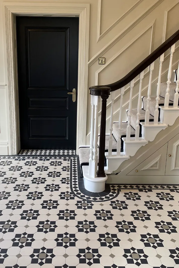 A view along the side of a grand staircase in a large entrance hall. The floor has Victorian floor tiles in black, white and grey tones. The border curves around the foot of the staircase. The staircase has cupboards underneath, and the walls have white wooden panelling. A black door leads to another room.