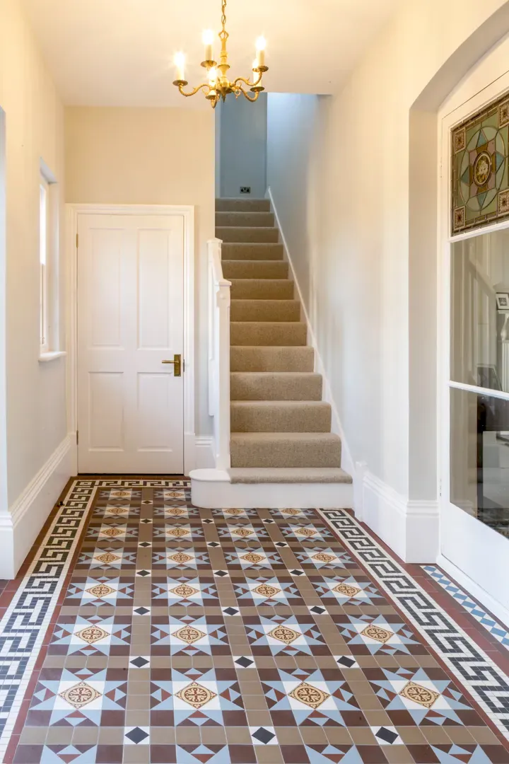 A wide hallway has neutral white paintwork throughout. To the rear a staircase with beige carpet rises, and a door with brass handle. The floor has a detailed multi-colour Victorian style mosaic tile pattern, with a Greek-key style border motif.