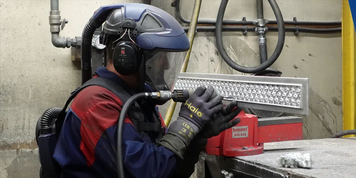 Man in a protective suit, gloves and helmet, hand finishes an aluminium daisy grate vent with a dremel tool.