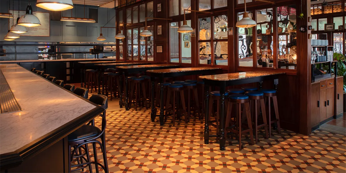 A restaurant with wooden panelling, and high tables with stools. The floor has an Edwardian style mosaic pattern in brown, yellow and red tones, based around an octagon shape. To the left and rear, the kitchen serving area with lamps hanging above.