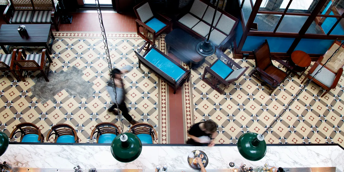 Overhead view of an Indian resturant bar area, with an Edwardian-type of mosaic floor pattern in yellow, red and brown tones. There are various seats and stools in different styles, and two large green lamps hang from the ceiling over the marble topped bar. Two blurred figures are seen moving through the space.