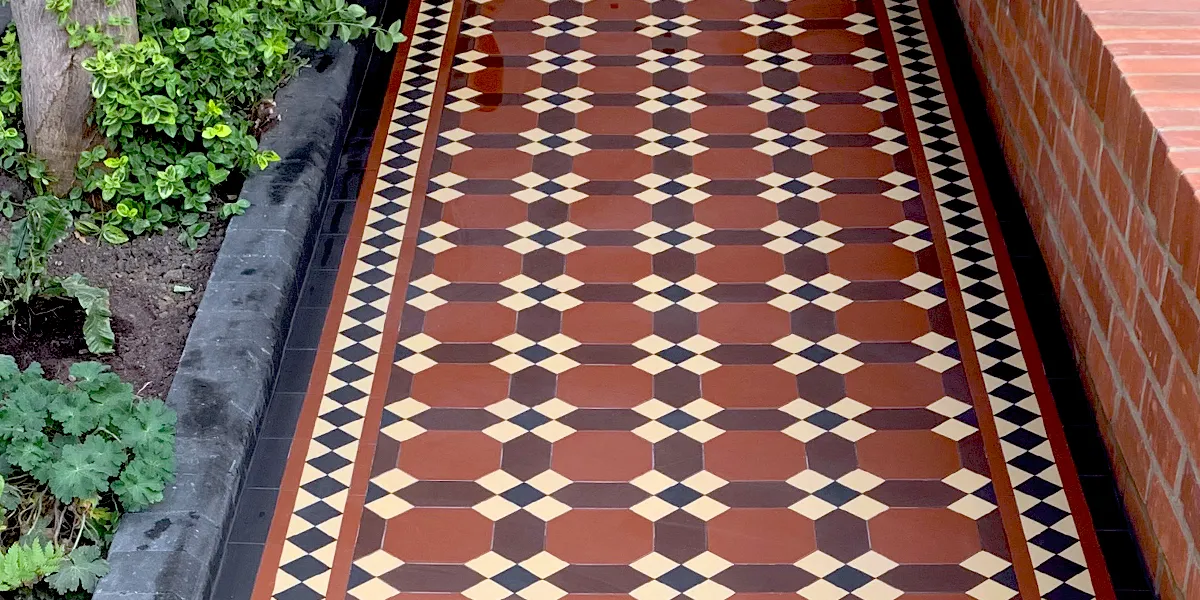 Red, brown and yellow mosaic tiled pathway with a red brick wall to the right and some green planting to the left.